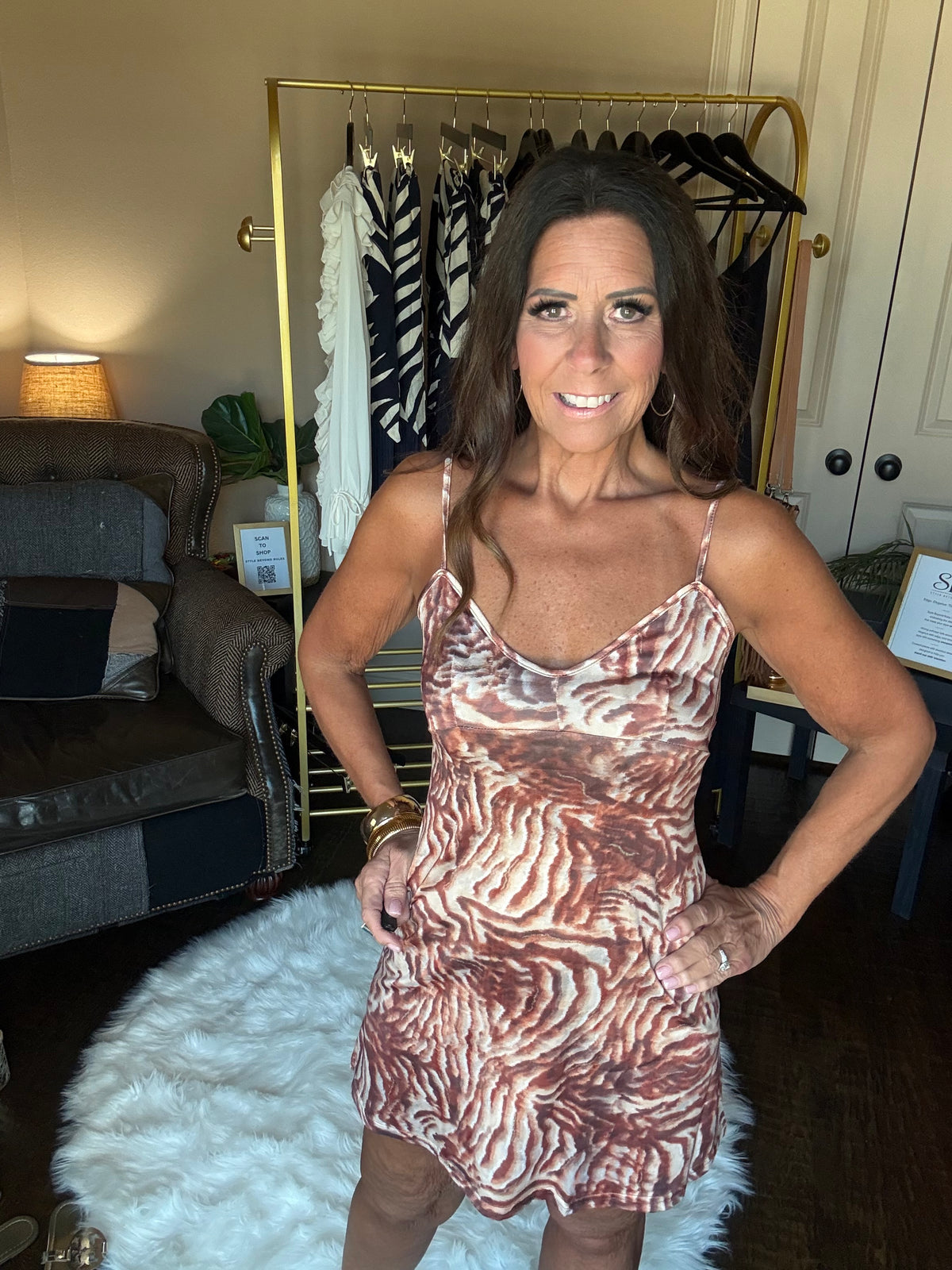 A woman wearing a brown and beige zebra-print spaghetti-strap dress, standing in a home setting with a clothing rack and sofa in the background.