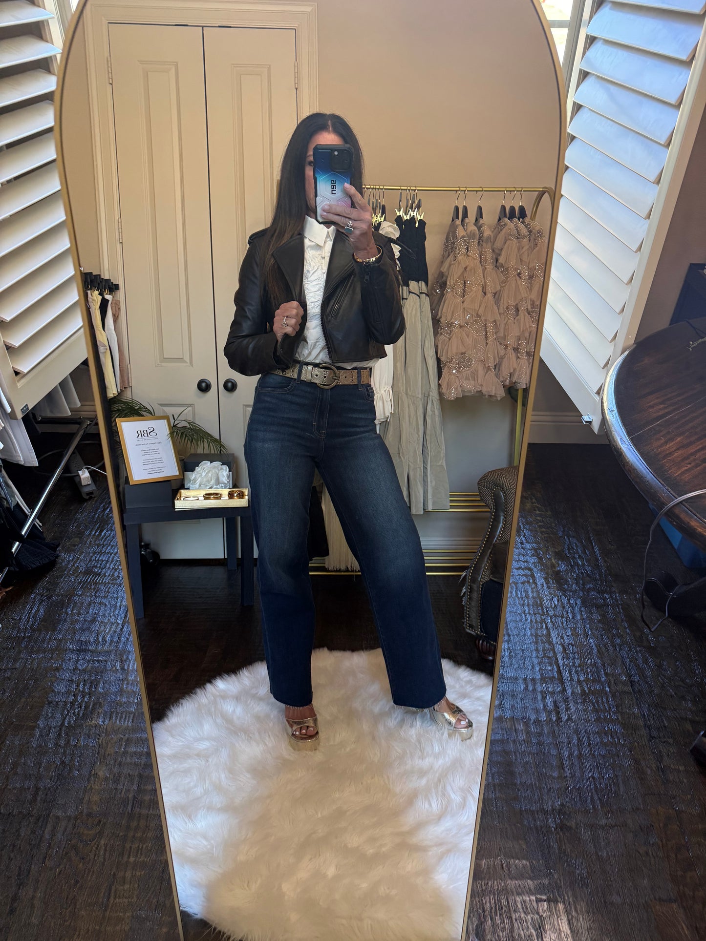 A woman takes a mirror selfie in a fitting room wearing a black leather jacket, white blouse, dark blue flare jeans, and metallic sandals. A clothing rack with garments and a white shag rug are visible in the background.