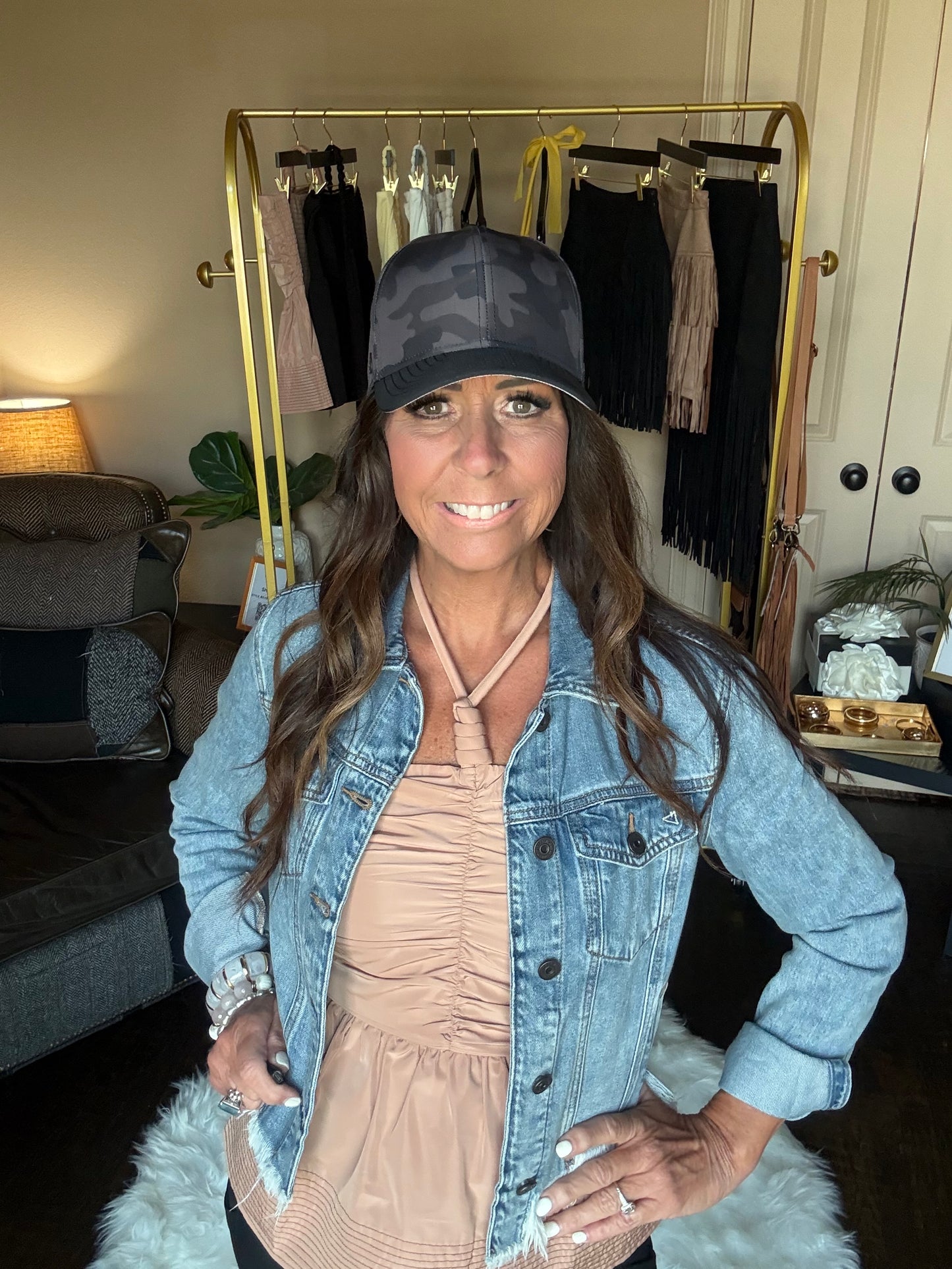 Woman wearing a denim jacket and black cap indoors with a mirror and clothing rack in the background.