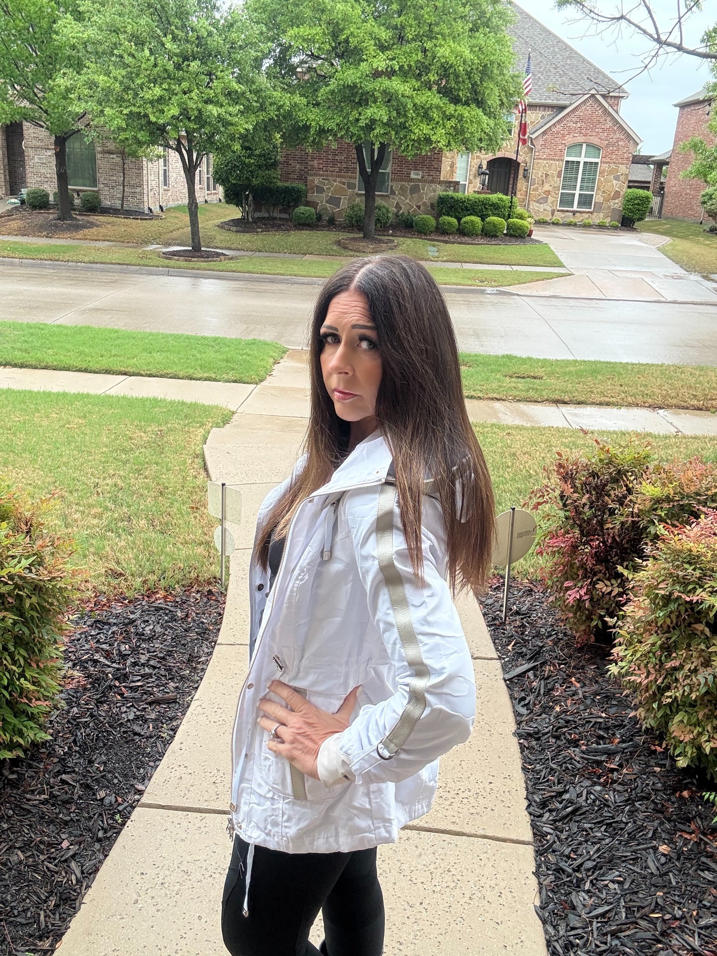 Woman in a white jacket standing on a sidewalk in a suburban neighborhood.