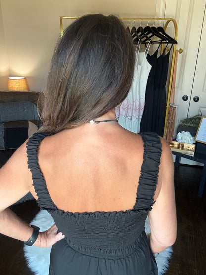 A woman viewed from behind wearing a black ruched spaghetti-strap dress, showing the back of the garment in an indoor room setting with a clothing rack visible in the background.