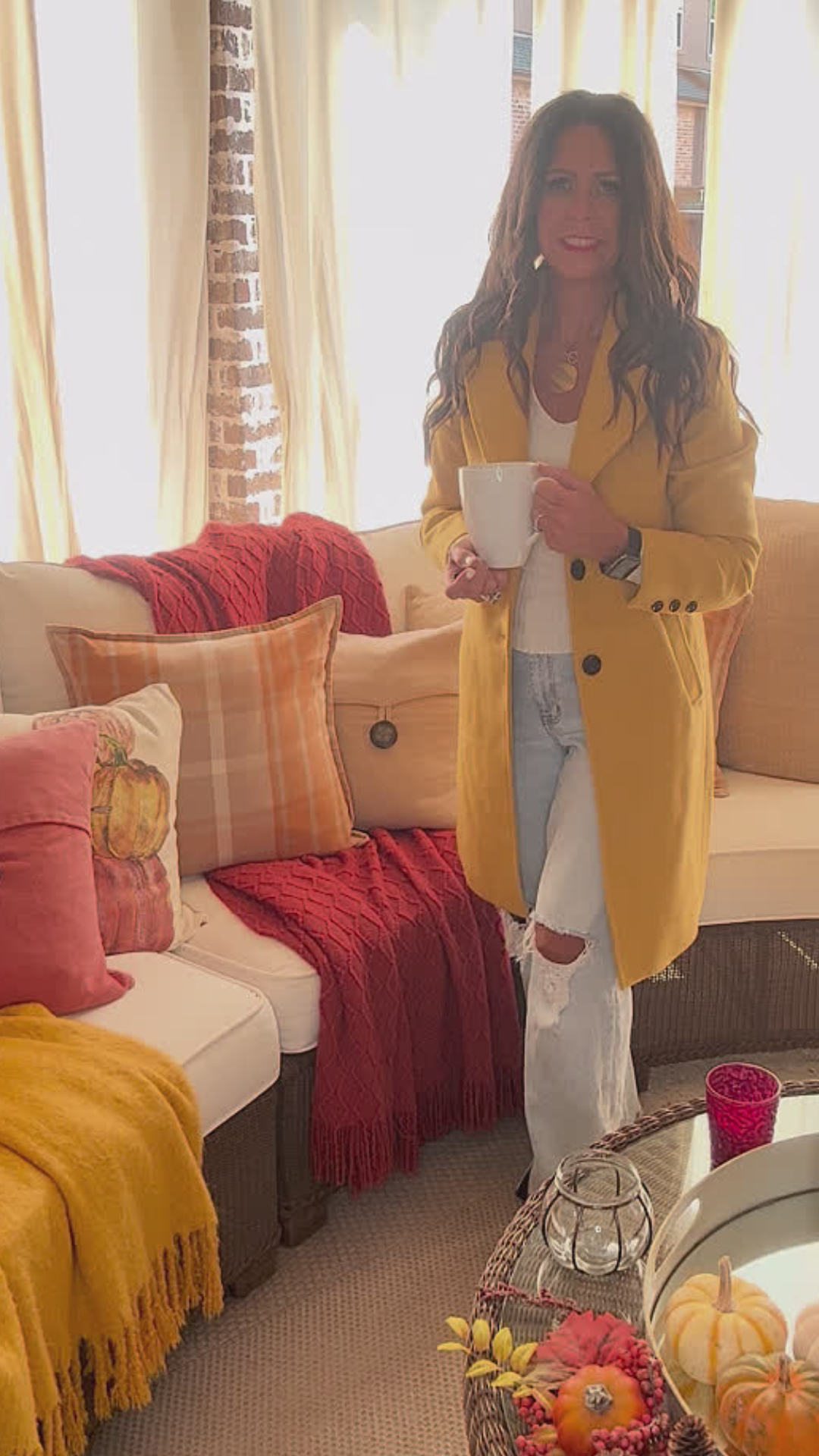 Model is wearing a yellow coat with a cream fitted top and distressed jeans while drinking coffee on a patio.