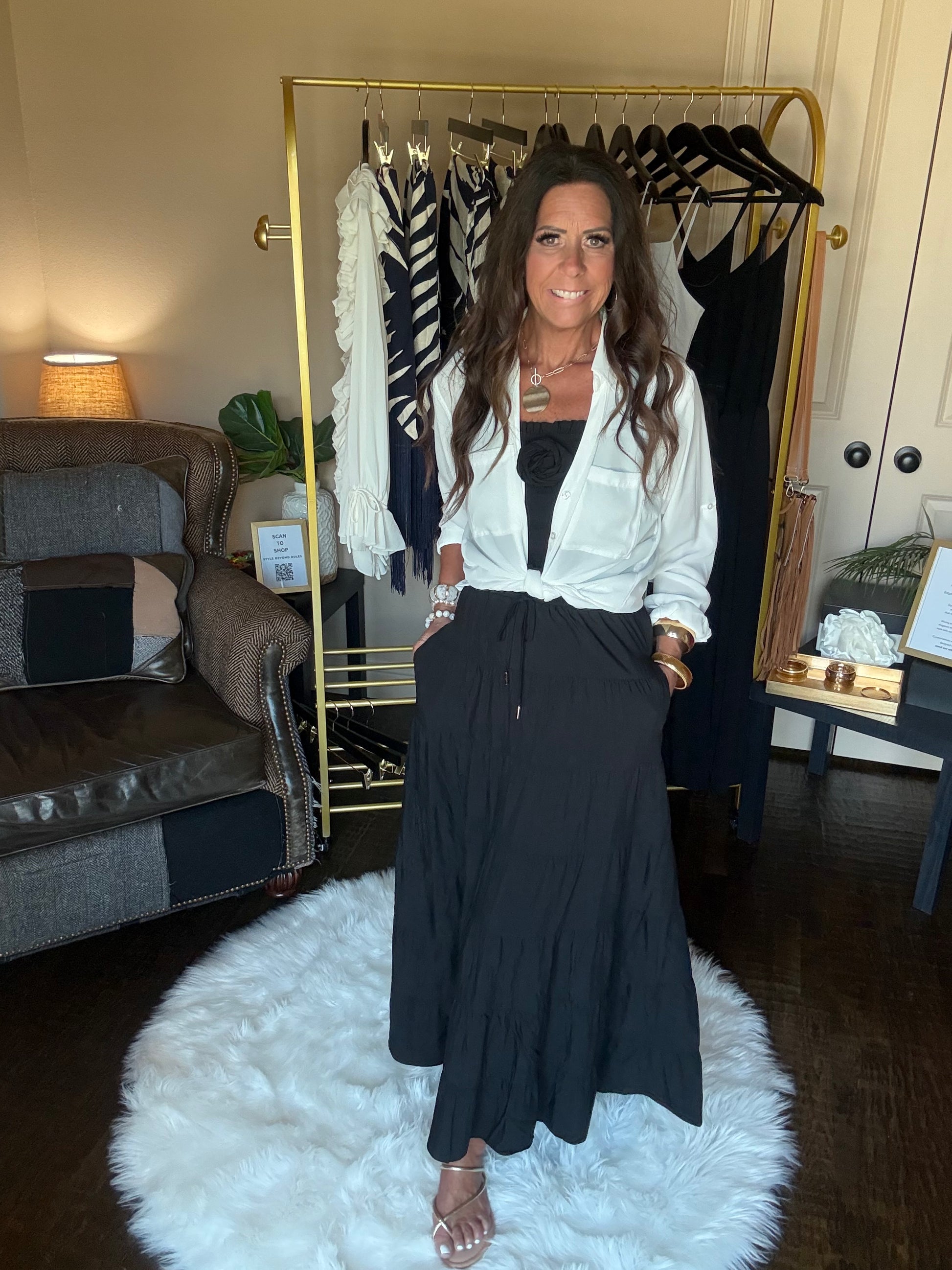A woman stands in a room wearing a white long-sleeve button-up shirt and a black pleated maxi skirt, with a gold clothing rack and a white fluffy rug in the background.