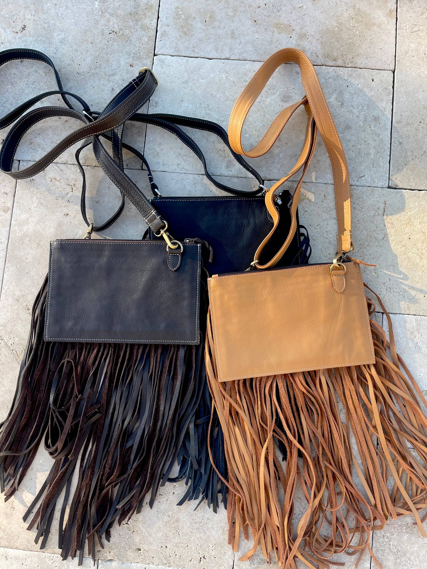Two handbags with fringe details, one black and one brown, on a stone surface.