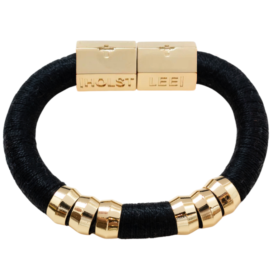 A black woven bracelet with gold-toned metal accents, featuring a box clasp engraved with the names 'HOLST' and 'LEE', and six cylindrical gold beads on the front.