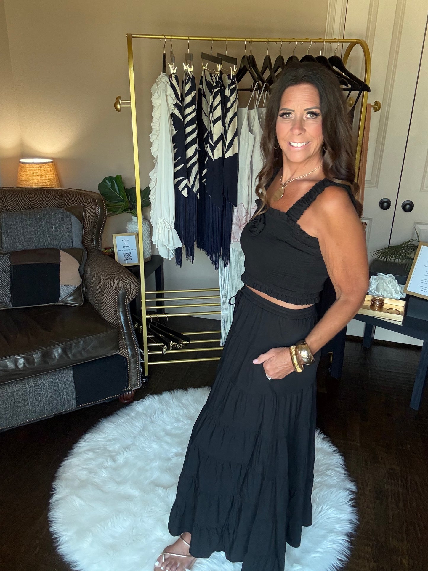 A woman models a black sleeveless maxi dress with a halter-style neckline and a matching pleated skirt in a home closet setting with a gold clothing rack and a white faux fur rug.