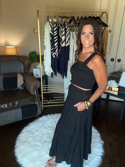A woman models a black sleeveless maxi dress with a halter-style neckline and a matching pleated skirt in a home closet setting with a gold clothing rack and a white faux fur rug.