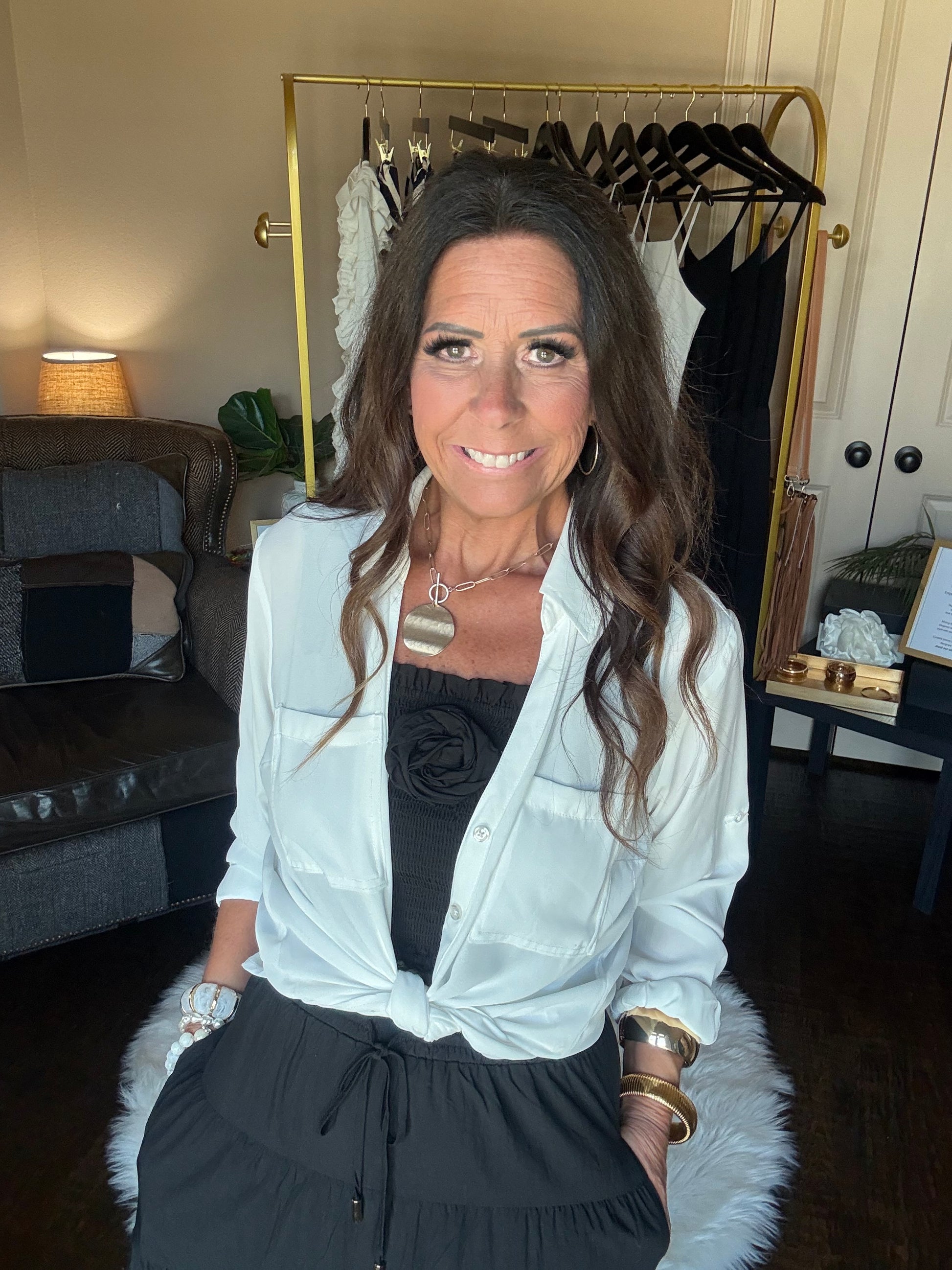 A woman wearing a white long-sleeve button-up shirt tied at the waist over a black top and black skirt, accessorized with gold bracelets and a silver pendant necklace, photographed in a home setting.