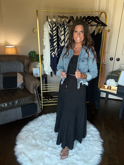 A woman stands in a room wearing a long black dress with a light blue denim jacket. A gold clothing rack with patterned shirts is visible behind her, along with a white fluffy rug, sofa, and dark wood floor.