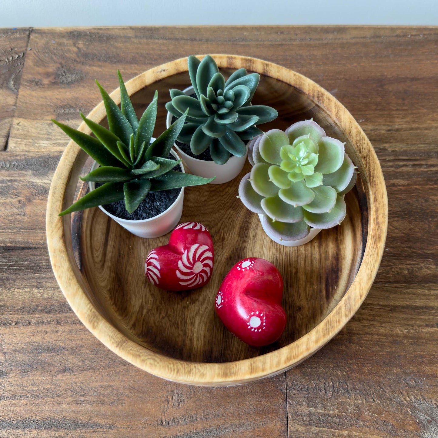 Three small potted succulents on a wooden plate with two red heart-shaped objects.