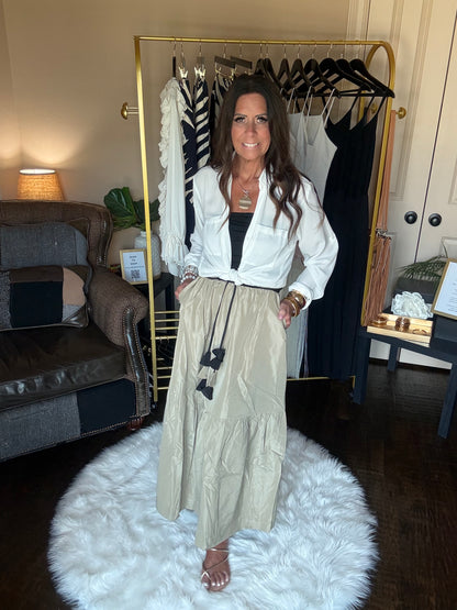 A woman models a white wrap-style blazer and beige wide-leg trousers with black bow accents, standing in a room with a clothing rack, sofa, and a white fluffy rug.