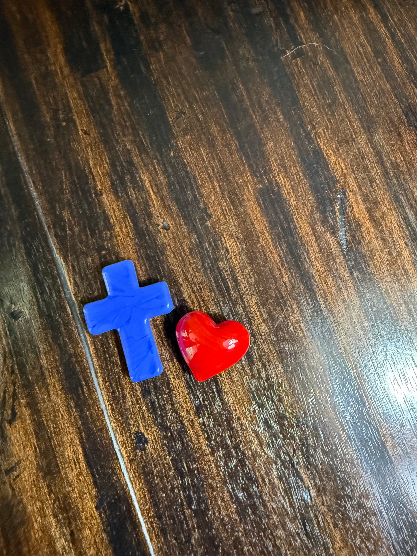 A small glossy red heart-shaped charm and a blue plus-shaped charm resting on a dark brown wooden surface.