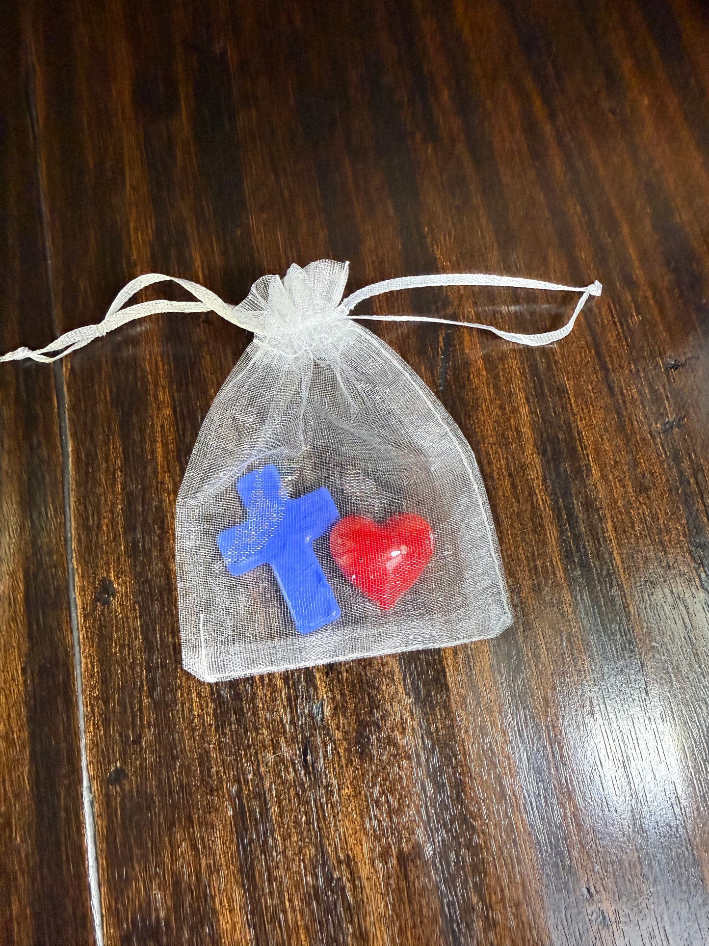 A small white organza drawstring pouch containing a red heart-shaped charm and a blue cross-shaped charm, placed on a dark wooden surface.