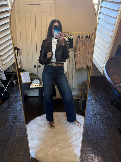 A woman takes a mirror selfie in a fitting room wearing a black leather jacket, white blouse, dark blue flare jeans, and metallic sandals. A clothing rack with garments and a white shag rug are visible in the background.
