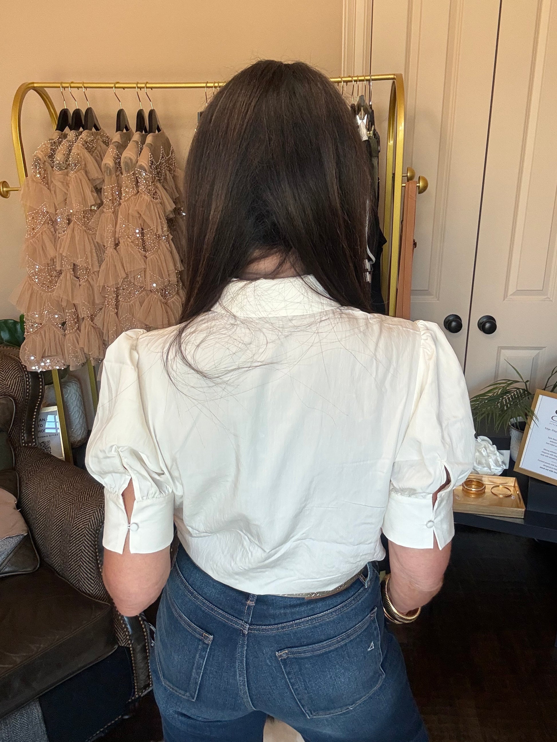 A woman viewed from behind wearing a white long-sleeve button-up shirt and blue jeans, standing in front of a clothing rack in an indoor setting.