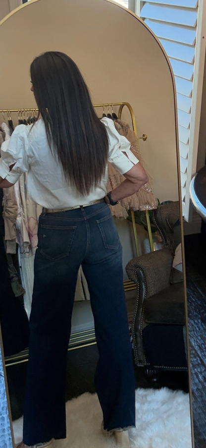 A woman stands in front of a mirror wearing a white ruffled short-sleeve blouse and dark blue wide-leg jeans in a boutique setting.