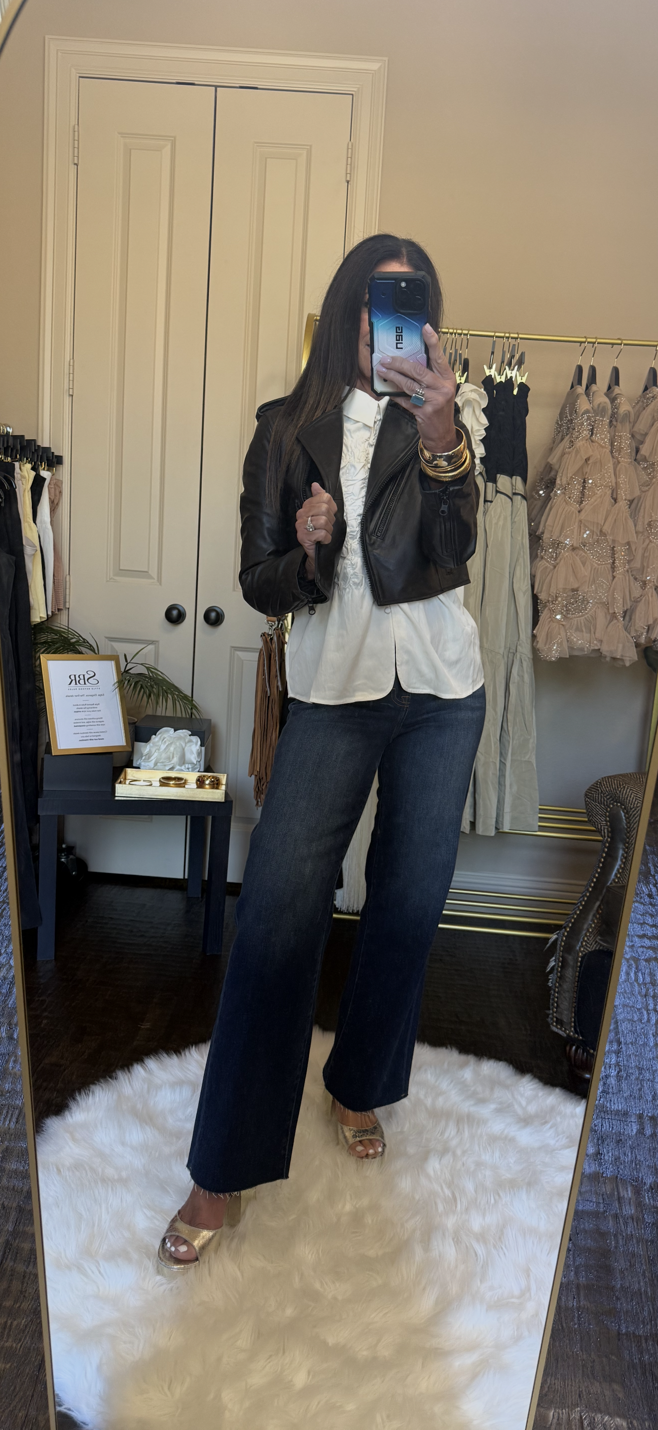 A woman takes a mirror selfie in a fitting room, wearing a black leather jacket, white button-down shirt, dark wide-leg jeans, and metallic heeled sandals.
