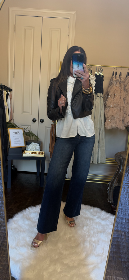 A woman takes a mirror selfie in a fitting room, wearing a black leather jacket, white button-down shirt, dark wide-leg jeans, and metallic heeled sandals.
