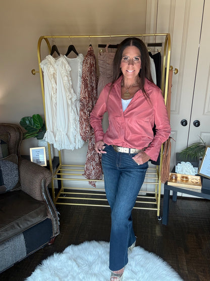 A woman stands in a home setting wearing a dusty pink satin button-down shirt and blue jeans, posing in front of a gold clothing rack.