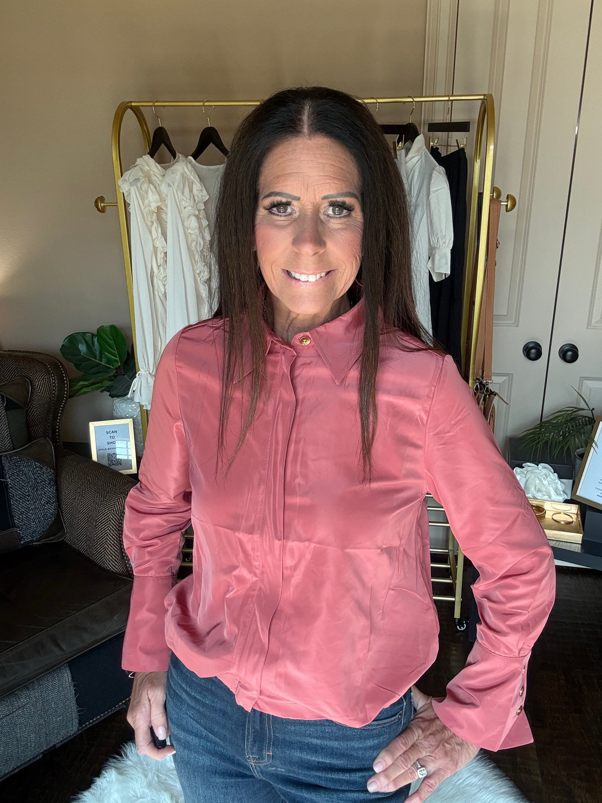 A woman wearing a dusty rose pink long-sleeve button-up shirt and blue jeans, standing in front of a gold clothing rack in an indoor room.