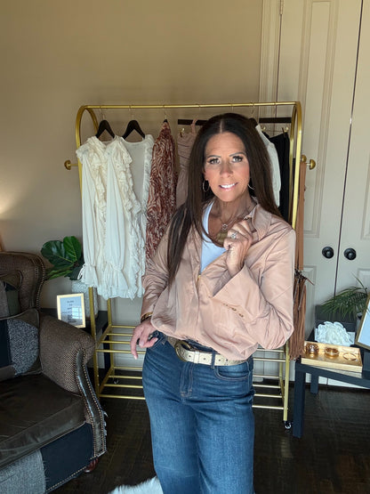 A woman stands in a room wearing a light beige button-up shirt and blue jeans, with a gold clothing rack behind her holding several tops.