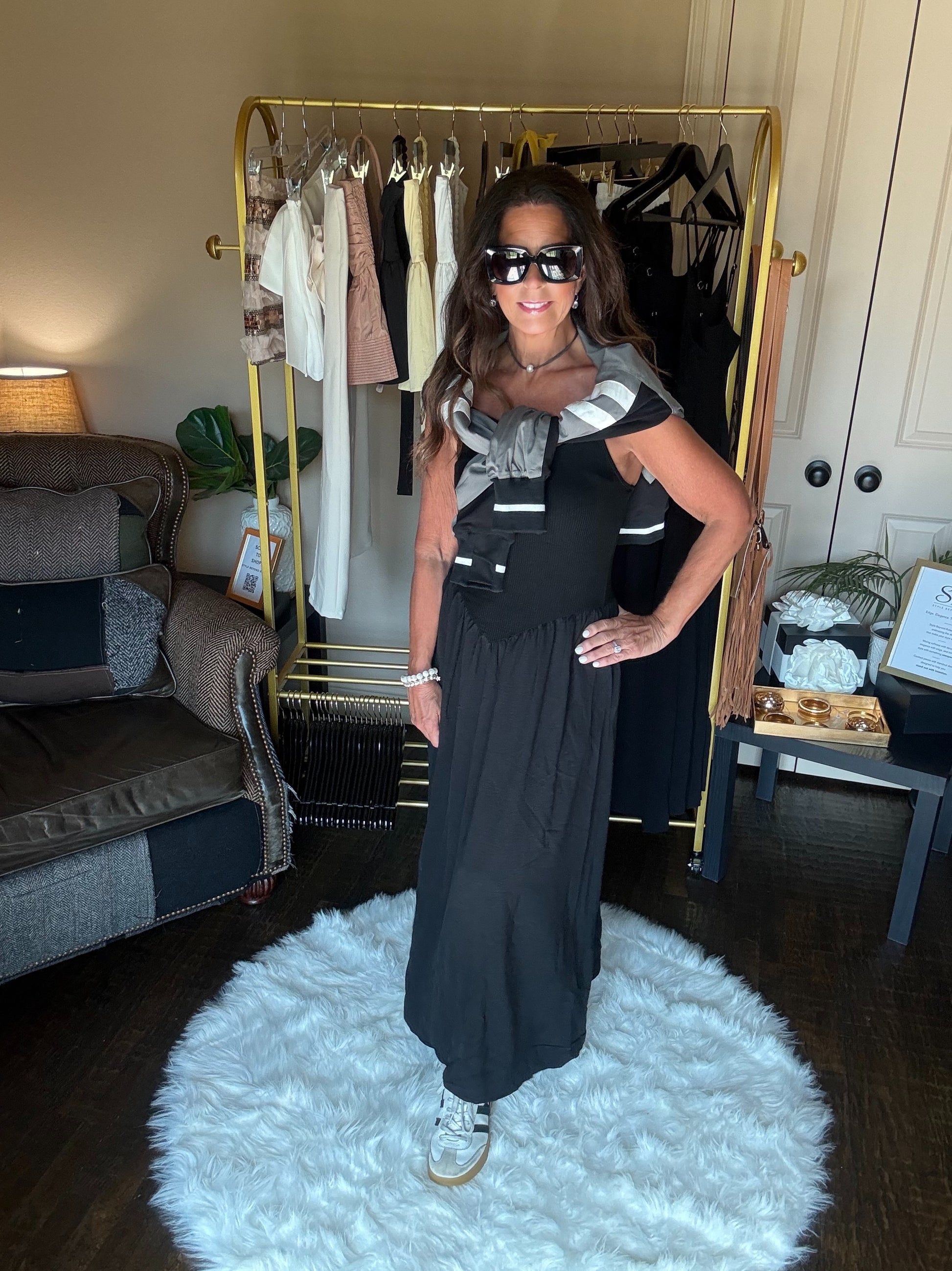 Woman in a black dress standing on a white rug in a room with a clothing rack and furniture.