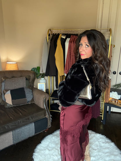 A woman stands in a room wearing a black faux fur cropped jacket and a burgundy satin skirt, holding a small metallic handbag. A clothing rack with various garments, a brown armchair, and a white fluffy rug are visible in the background.