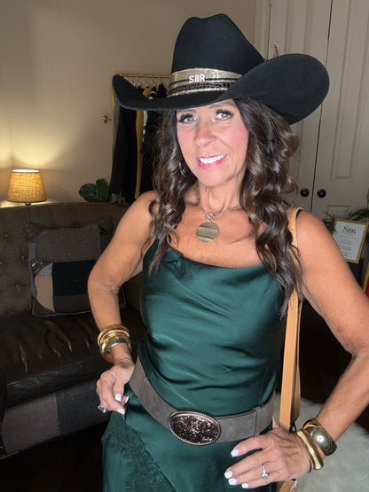 Model is wearing a hunter green satin and lace maxi dress with spaghetti straps and an asymmetrical hem and a cowboy chic vibe.