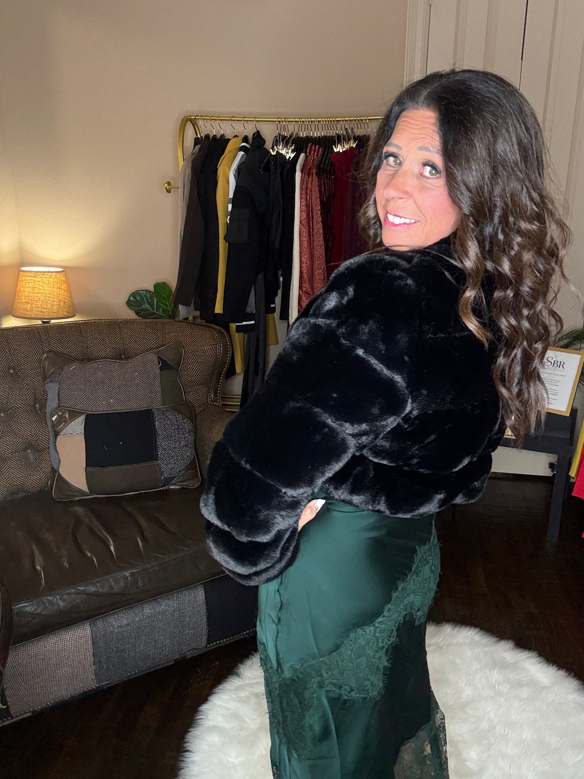 Model is wearing a hunter green satin and lace maxi dress with spaghetti straps and an asymmetrical hem layered with a black faux fur bolero jacket.