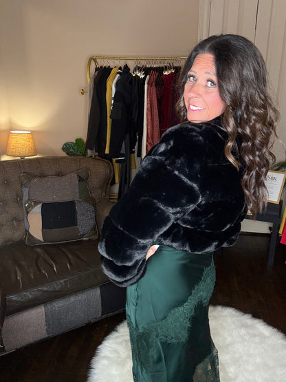 Model is wearing a hunter green satin and lace maxi dress with spaghetti straps and an asymmetrical hem layered with a black faux fur bolero jacket.