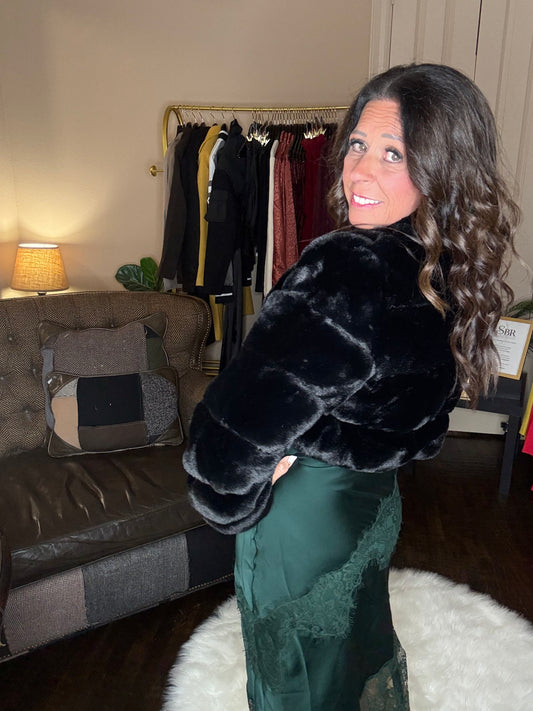 Model is wearing a hunter green satin and lace maxi dress with spaghetti straps and an asymmetrical hem layered with a black faux fur bolero jacket.