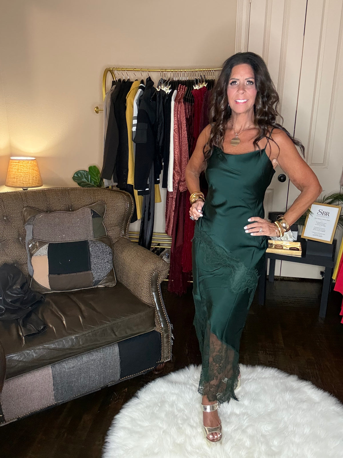 Model is wearing a hunter green satin and lace maxi dress with spaghetti straps and an asymmetrical hem land gold metallic heels.