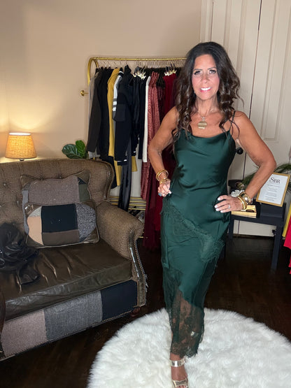 Model is wearing a hunter green satin and lace maxi dress with spaghetti straps and an asymmetrical hem land gold metallic heels.