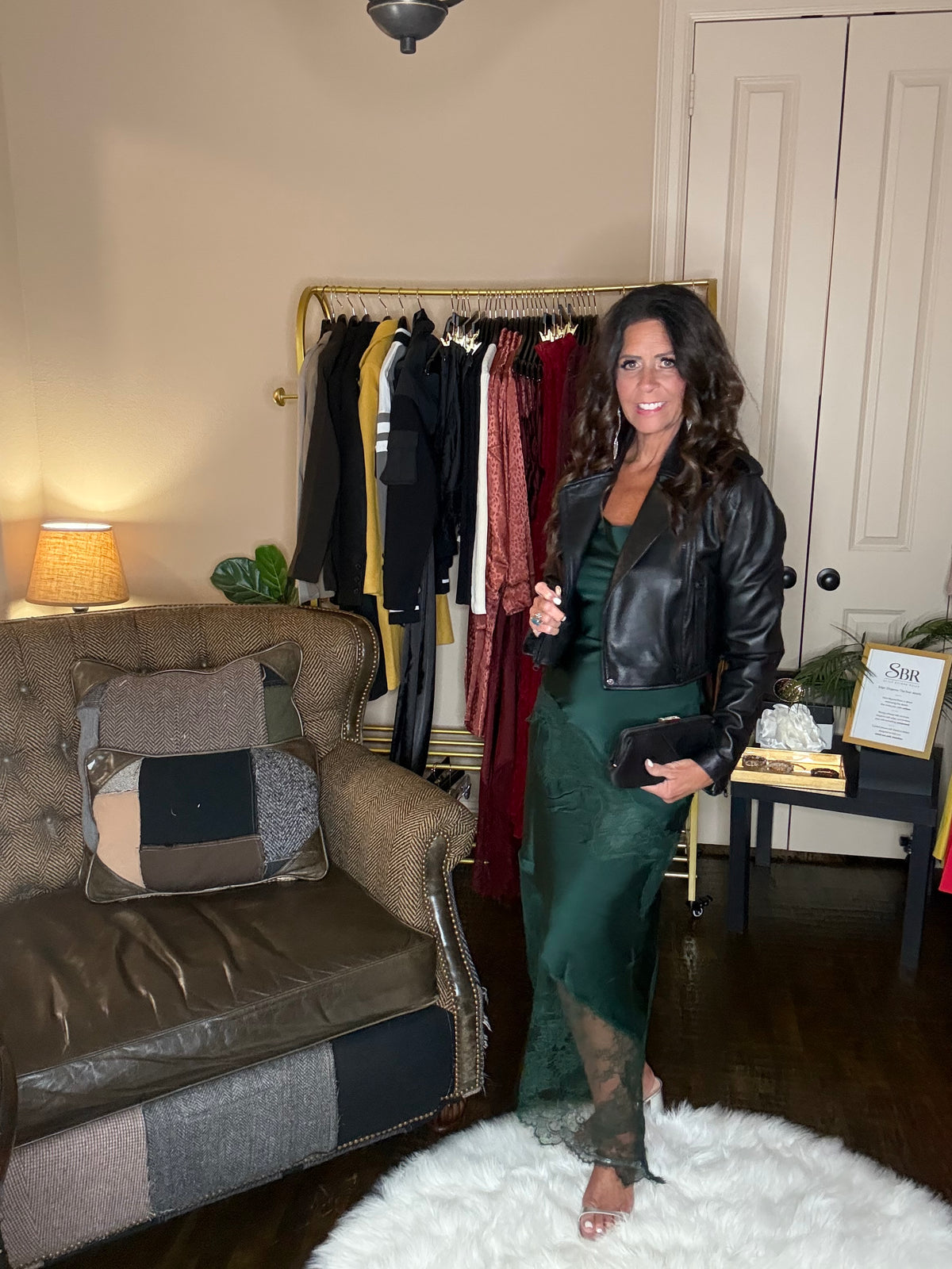 Model is wearing a hunter green satin and lace maxi dress with spaghetti straps and an asymmetrical hem land gold metallic heels and a blak cropped leather jacket.