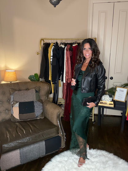 Model is wearing a hunter green satin and lace maxi dress with spaghetti straps and an asymmetrical hem land gold metallic heels and a blak cropped leather jacket.