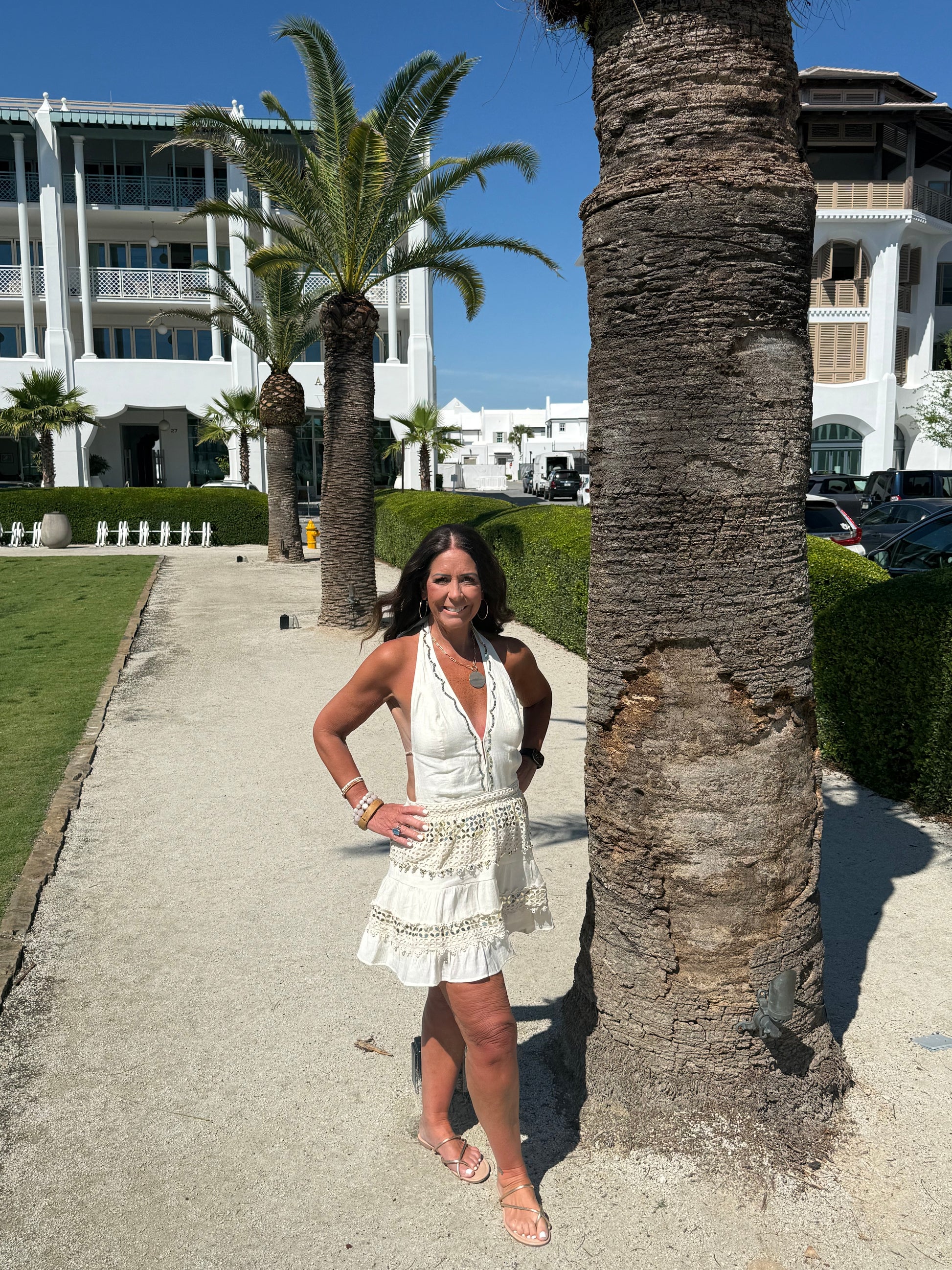 woman posing by a palm tree wearing a cream and gold halter dress
