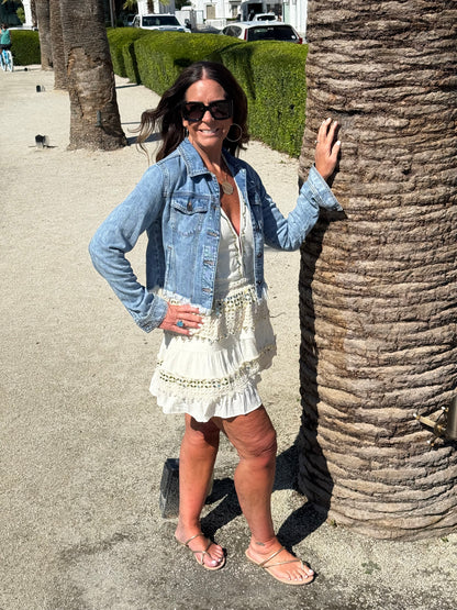 woman posing by a palm tree wearing a cream and gold halter dress layered with a cropped jean jacket