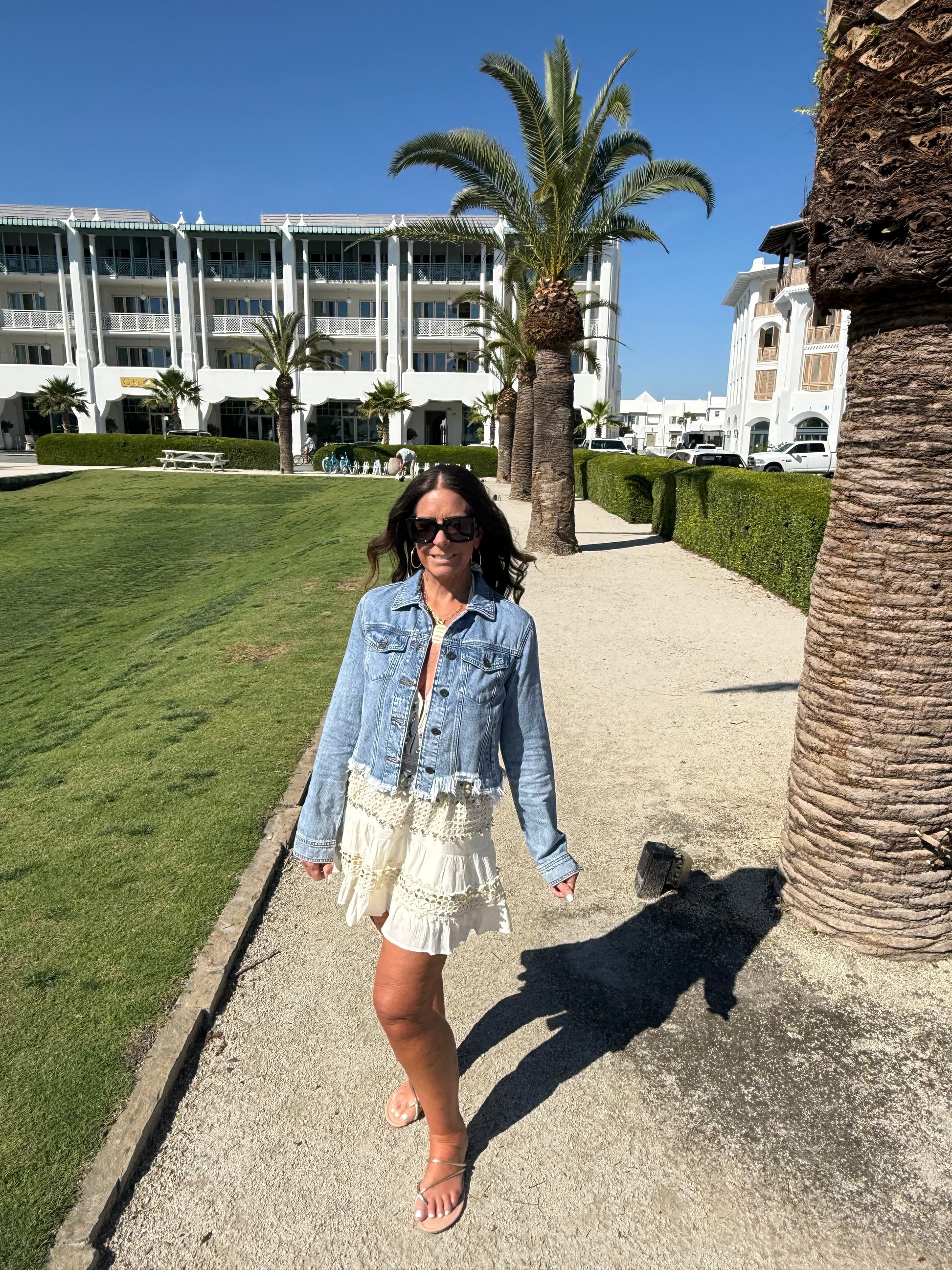 woman posing by a palm tree wearing a cream and gold halter dress layered with a cropped jean jacket