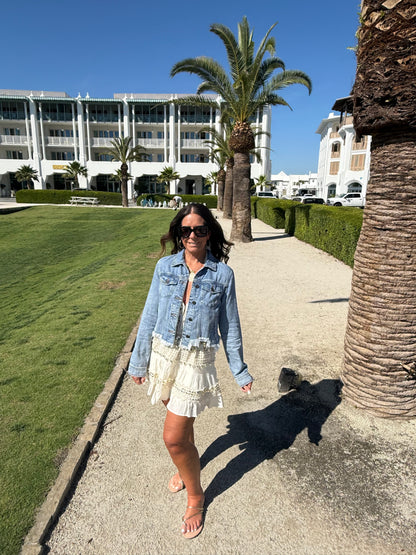 woman posing by a palm tree wearing a cream and gold halter dress layered with a cropped jean jacket