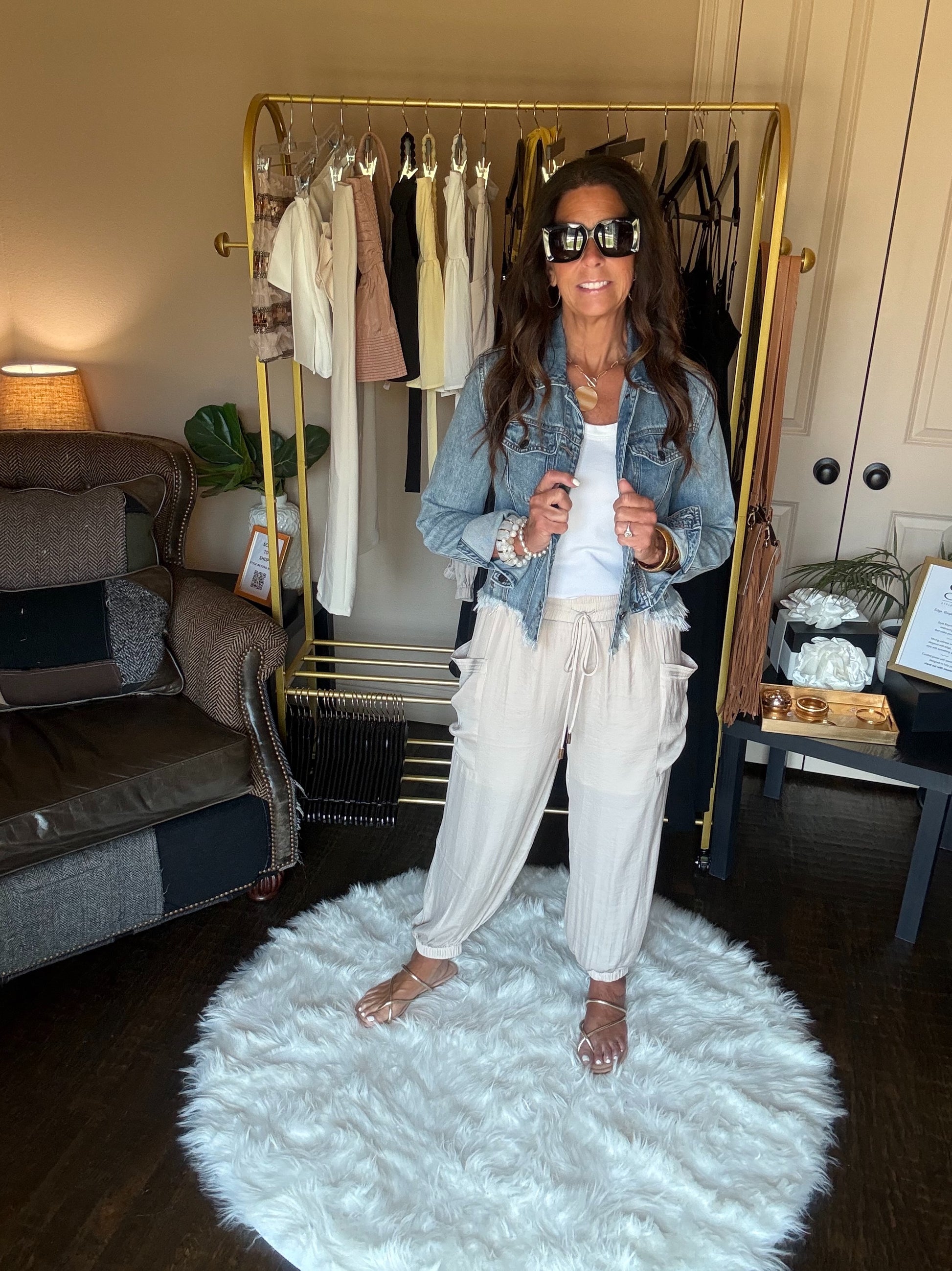 Woman standing in a room with a rack of clothes, wearing a denim jacket and light pants.
