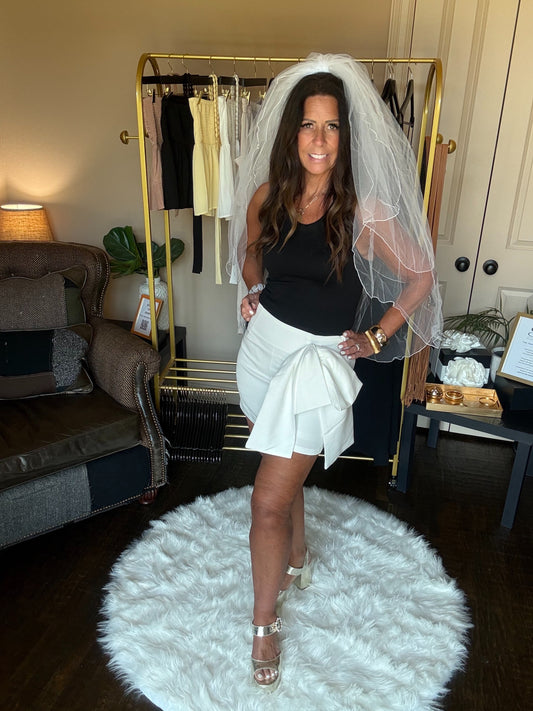 Woman in a black top and white skirt with a veil standing on a white rug in a room with a clothing rack and furniture.