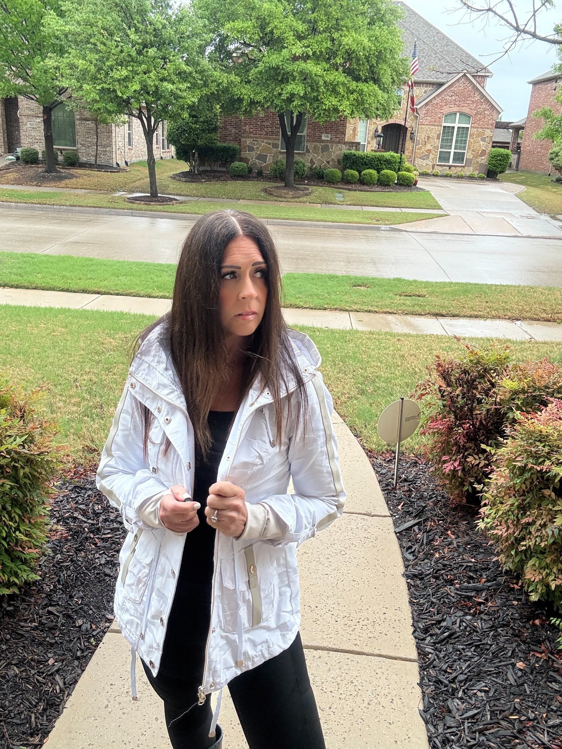 Woman in a white jacket standing on a sidewalk in a suburban neighborhood.