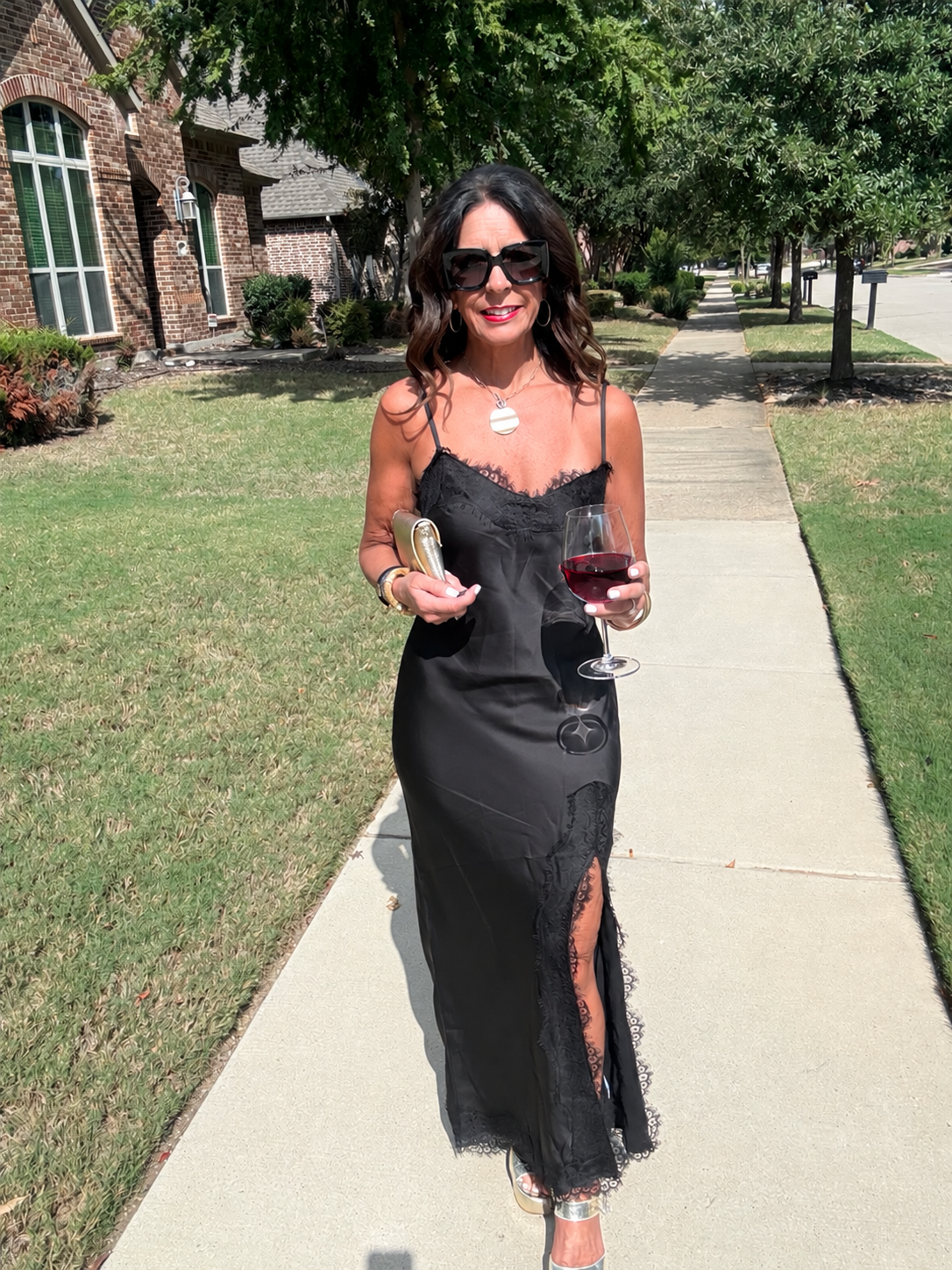 Black satin lace trim slip maxi dress styled with a metallic clutch and heels, front view holding a wine glass.