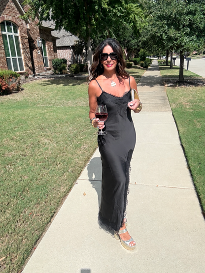 Black satin lace trim maxi dress styled for evening with a metallic clutch, platform heels and wine glass.