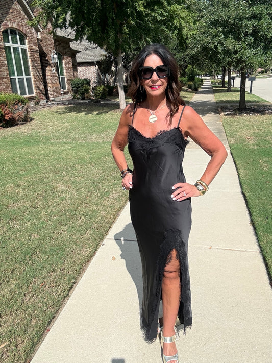 Front view of black satin lace trim slip maxi dress with spaghetti straps, lace neckline and high slit, styled with metallic heels.