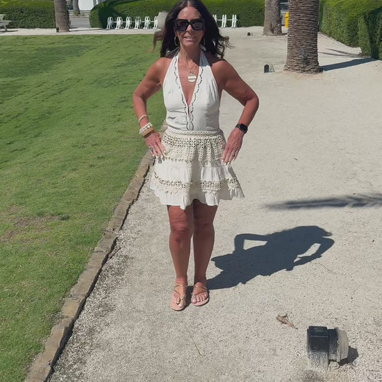 Woman walking on a path with white buildings and palm trees in the background and she is modeling a cream halter dress  with gold accents