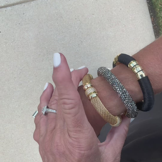 A black woven bracelet with gold-toned metal accents, featuring a box clasp engraved with the names 'HOLST' and 'LEE', and six cylindrical gold beads on the front paired with two other bracelets as a bracelet stack.
