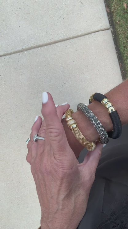 A black woven bracelet with gold-toned metal accents, featuring a box clasp engraved with the names 'HOLST' and 'LEE', and six cylindrical gold beads on the front paired with two other bracelets as a bracelet stack.