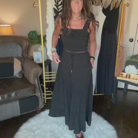 A woman viewed from behind wearing a black ruched spaghetti-strap dress, showing the back of the garment in an indoor room setting with a clothing rack visible in the background.