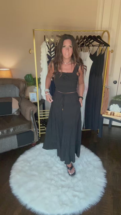 A woman viewed from behind wearing a black ruched spaghetti-strap dress, showing the back of the garment in an indoor room setting with a clothing rack visible in the background.